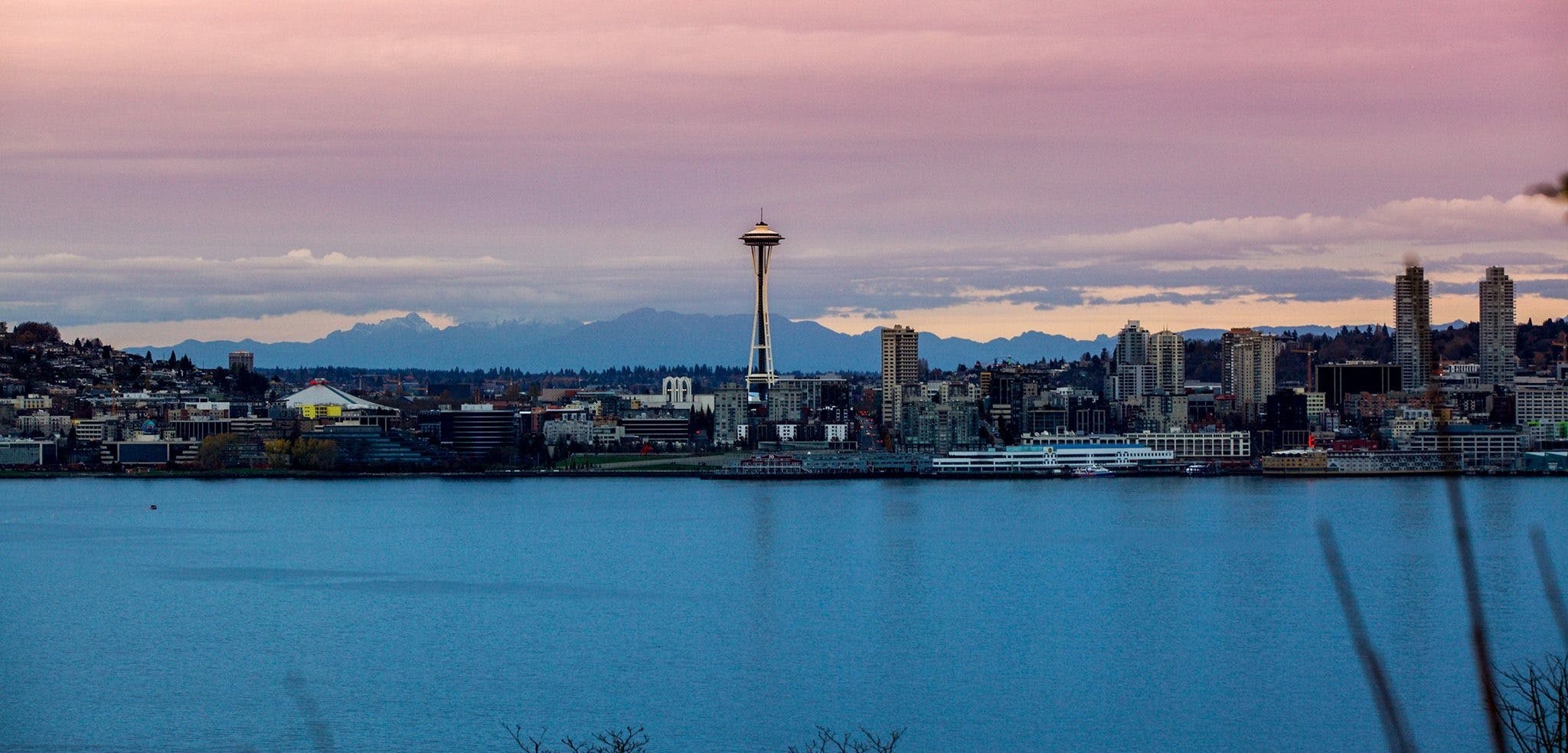Seattle skyline with the Space Needle, set against a sunset sky and a body of water in the foreground.